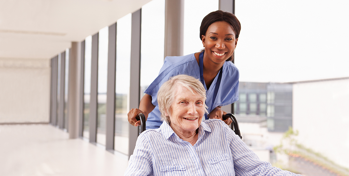 Nursing Assistant Pushing Senior Patient In Wheelchair Along Corridor